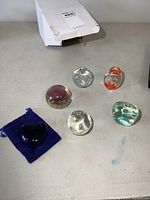Full view of all six paper weights, including the dark blue heart resin weight on a blue pouch and five glass paper weights with various internal designs.