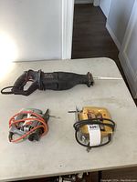 Three power saws on white table: one large black reciprocating saw and two Black & Decker jigsaws with power cords.