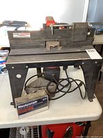 Front view of the Sears Craftsman router table showing mounted router underneath, power cord, and small plastic box of router bits beside it.