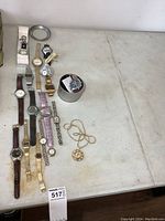 Overhead view of 13 assorted mens and ladies wristwatches, including digital watch with cracked screen, some with metal bands, some leather, and a Hersheys branded watch. Also visible a pendant watch necklace.