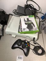 White Xbox 360 console with black controller, power supply, and 4 game cases stacked including Call of Duty Modern Warfare 3