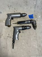 Three air drills laid out on a surface: one straight with chuck, one straight smaller drill, one right-angle drill with chuck.