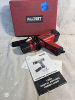 Red Alltest 1100A digital timing light with wires, original red hard case and instruction manual laid out on a white surface.