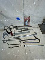 Photo showing various automotive repair hand tools on the ground including pry bars, large ring wrenches, and a tool in unopened packaging.