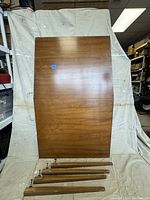 Full view of the rectangular teak tabletop leaning against a backdrop with five detachable legs laid in front of it on the floor.