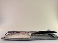 Full view of a rectangular silver-plated crumb tray with an engraved surface pattern and a textured dark handle, shown resting on a gray felt pouch.