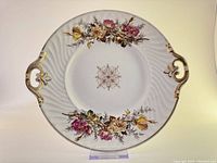 Photo of the antique porcelain serving dish showing the floral design, gold-tone handles and rim, embossed swirl border, and central decorative motif.