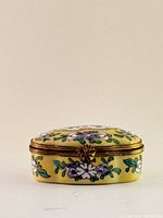 Front view of oval porcelain trinket box showing yellow background and hand-painted purple and white flowers with green leaves, gold-tone trim and floral clasp.