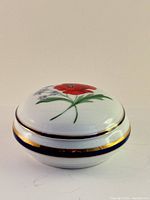 Side view of the round porcelain trinket box showing the domed lid with hand-painted red poppy and navy blue and gold bands around the edge.