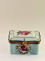Front view of pale green rectangular porcelain trinket box with hand-painted large pink rose and gold accents on lid and front side, showing gold-tone clasp.