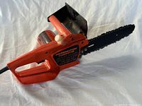 Right side view of Remington Powercutter electric chainsaw, showing orange plastic body, black blade, white adjustment knob, and partial view of motor housing and cord.
