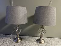 Pair of modern table lamps on gray countertop with gray wall background. Each lamp has a brushed nickel spiral openwork base and matching gray fabric drum shade. Both lamps are shown with cords visible. Lighting highlights brushed metal finish and fabric textures.