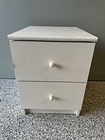Front view of the IKEA white nightstand showing two drawers with round white knobs and visible wear on surface.