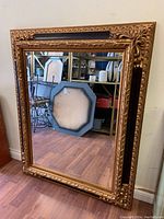 Full view of the large rectangular wall mirror with gold and black ornate frame leaning against the wall, showing the overall condition and size.