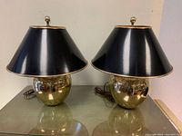 Pair of gold toned metal table lamps with black lampshades, placed side by side on glass surface.