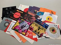 Top-down photo showing a pile of over 25 assorted 7-inch vinyl records in sleeves, some loose vinyl visible