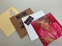 Four vintage rock vinyl record albums stacked showing the fronts and spines including Rod Stewart Greatest Hits, AC/DC, The Clash, and The Who albums.