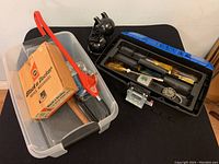 Opaque plastic bin with Black & Decker drill box, metal hammer, tubing, and a small black desk lamp. Also part of a blue toolbox with assorted tools visible in the top tray.