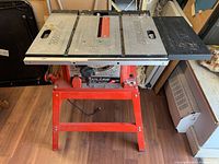 Front view of Skilsaw table saw on red stand on wooden floor, showing the table surface with Skilsaw branding and blade slot.