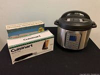 Photo of the three appliances: an InstaPot pressure cooker, boxed Cuisinart electric knife with stand, and boxed ultrasonic vacuum bag sealer, arranged on a surface against a plain wall.