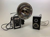 Full view of Kodak Duaflex III camera with mounted Kodalite Flasholder Lumaclad reflector and nearby Clix-O-Flex camera on white background.