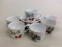Photo showing six mugs with fruit designs (cherry and grape) and six matching saucers.