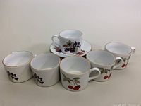 Photo showing six white porcelain mugs, five decorated with red cherries and one with grapes, all with gold rim