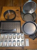 Multiple baking pans including round cake pans, a glass pie plate, madeleine mold pan, and silicone brush arranged on a wooden surface.