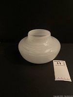 Full view of a white blown glass vase with subtle white thread-like swirls, round body, short neck, and flared rim on a black background.