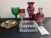 Photo showing full lot of colored glassware including candy dish, goblets, vase, butter dish, and bottles arranged on black surface