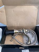 Top view of single lever bath faucet inside packaging box showing chrome finish, attached hose, and black mounting base.