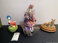 Three musical figurines: a goose in bonnet and red scarf, a clown figure with purple and pink clothes missing one hand, and two rabbits on a wooden base.
