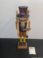 Full front view of the nutcracker showing the entire figure, including its wooden base, top hat decorated with a flower, and wooden tool on the shoulder.