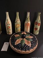 Photo showing four tall decorative bottles hand-painted with fruit and vegetable motifs behind a large ceramic pie plate with lid.