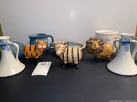 Front view of novelty animal pottery mugs (tiger, giraffe, lion) and pair of white candle holders with blue ribbons, and a white pot in the background.