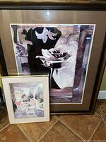 Two framed prints leaning against a wall on the floor; one large featuring a white rose and abstract architectural elements in purples and beige, the other smaller showing a porch scene with wicker chairs and flowers.