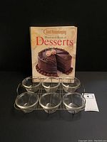 Set of six clear Pyrex custard dishes in metal rack with Good Housekeeping dessert cookbook behind, on black background