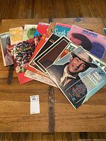Photo showing 11 vintage vinyl records spread out on a wooden floor with visible titles and cover imagery.