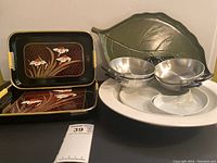 Two rectangular serving trays with floral design and yellow handles next to a green leaf-shaped serving tray, two metal egg skillets with black handles, and two white serving bowls