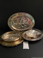 Four silverplate serving trays arranged against a black background showing different shapes, sizes, and designs including leaf pattern and scalloped edge.