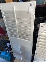 Front view of one white wood shutter showing horizontal slats and hinge hardware with other shutter partially visible.