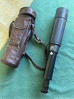 Bisley Deluxe scope with rubberized eyepiece and adjustment knobs, next to brown leather carrying case with shoulder strap and front pockets, all placed on a green tablecloth.