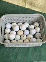 Top-down view of 25 mixed brand golf balls in a gray plastic basket on a green tablecloth showing mostly white balls with some variety of markings.