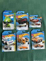 Six Hot Wheels toy cars in original packaging arranged on green surface: Angry Birds green pig car, Batman Batmobile, red and white sports car, silver and white offroad truck, orange race car, brown vintage car.