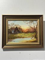 Frontal view of oil painting showing pastoral scene with sheep, trees, house, pond, and sunset. Features gold and brown frame and visible signature.
