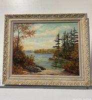 Full front view of oil painting depicting a lake scene with trees, rocks and sky in ornate white frame