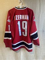 Back view of red hockey jersey with YZERMAN lettering and number 19, black and white stripes on sleeves and hem, hanging on white hanger.