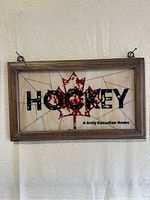 Front view of the vintage hockey glass sign showing the word 'HOCKEY' on a red maple leaf with black text reading 'A truly Canadian Game'. The wood frame and metal hooks for hanging are visible.