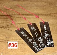 The photo shows three rectangular silver-plated Asian bookmarks with red cords. Each bookmark is engraved with Asian calligraphy and floral images including flowers and bamboo, placed on a wooden surface.