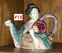 Front and angled view of the figurine teapot depicting a Japanese woman with teacup and fan, set against a wood background.